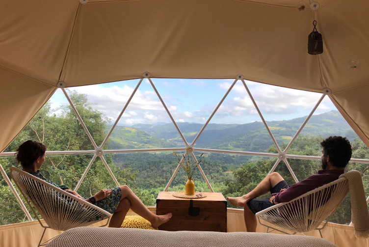 a man and a woman look at the view of the forest from glamping geodesic dome through panoramic window