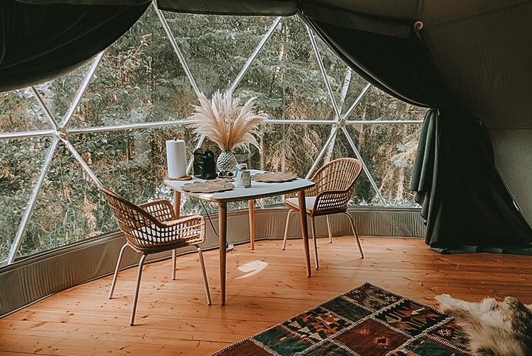 the interior of a geodesic dome with a view of the window, chairs and a coffee table, and the forest outside the window