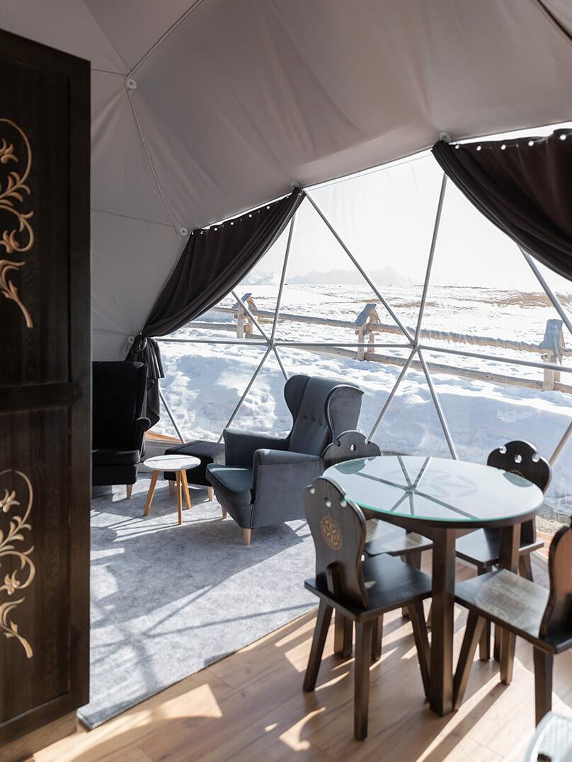 interior of a geodesic dome with a view of armchairs, coffee table, chairs and dining table, snow outside the window