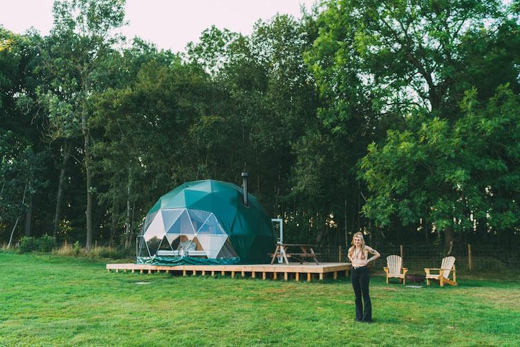 woman in front of a forest green geodesic dome in the forest