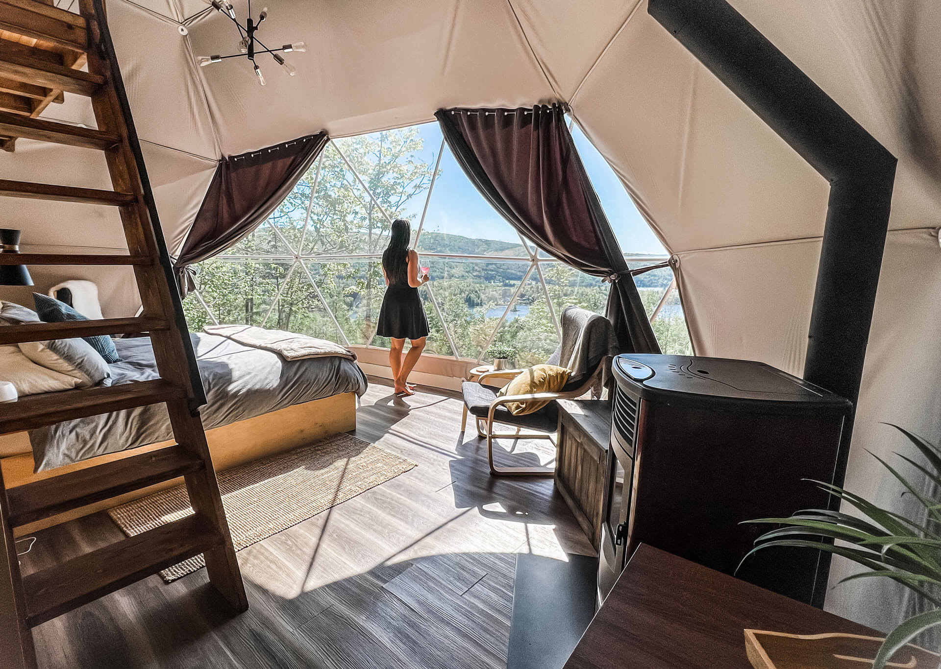 woman standing in front of windows inside of geodesic dome with a bed, a stove an armchair
